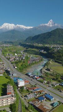 Aerial portrait vertical drone shot of a village in Pokhara beside Prithvi Highway near dense forest and the Gandaki River with snow-capped Machhapuchhre and Annapurna mountain range in Nepal.