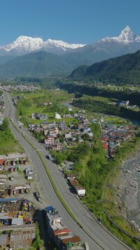 Aerial 9:16 Vertical shot of Prithvi Highway Pokhara Nepal passing a colorful settlement near the blue Gandaki River with dense forest and Machhapuchhre and Annapurna peaks rising.