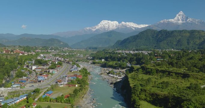 Drone shot of Pokhara Nepal with colorful rooftops along the Prithvi Highway beside the Gandaki River surrounded by dense green forest with Annapurna and Machhapuchre Himalayan peaks.