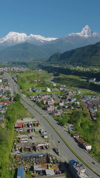 Aerial 9:16 Portrait drone shot of Prithvi Highway Nepal Pokhara running through a colorful settlement beside the blue Gandaki River with green dense forest and Annapurna and Machhapuchhre peaks