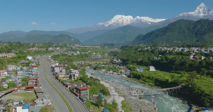 Aerial shot of rural Pokhara Nepal with Prithvi Highway running beside the blue Gandaki River through green fields and forest with Annapurna and Machhapuchhre peaks rising.