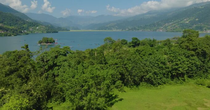 Aerial shot pulling into Tal Barahi Temple Pokhara Nepal on an island in clear blue Phewa Lake surrounded by green forest under a cloudy blue sky.