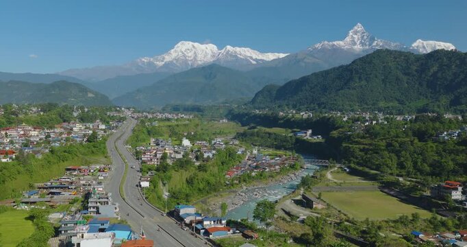 Aerial shot of Prithvi Highway passing a colorful village and green forest beside the Gandaki River with Annapurna and Machhapuchhre peaks under a clear blue sky in Pokhara Nepal.