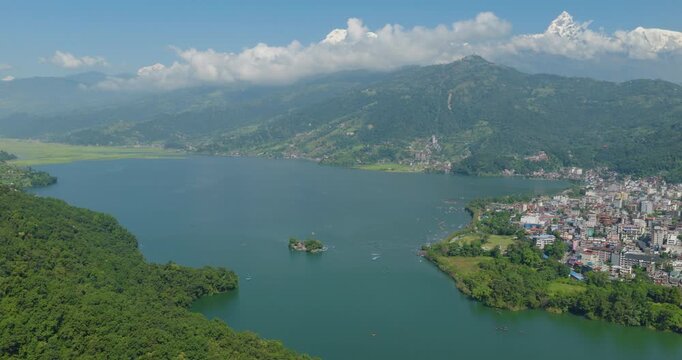 Aerial shot of calm blue Phewa Lake surrounded by green forest with Annapurna and Machhapuchhre peaks rising beneath a cloudy blue sky in Pokhara Nepal.