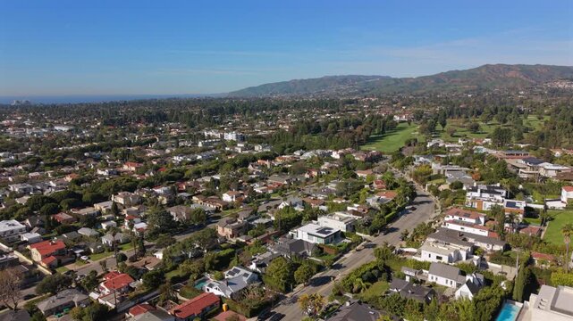 Aerial los angeles suburb panoramic view showing sunlit residential streets, treelined yards, modern houses, golf green in distance, rolling hills and distant coastline under clear blue sky,
