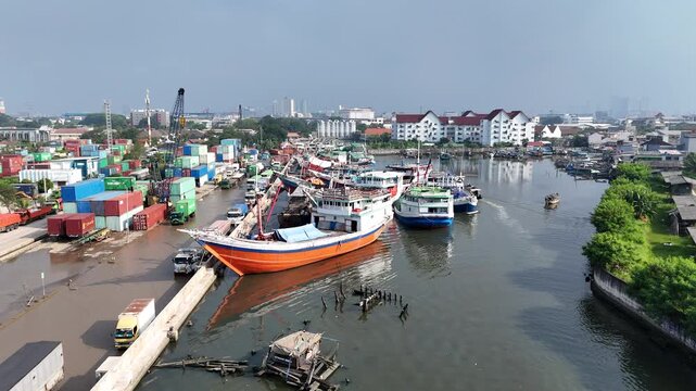 Aerial view of traditional wooden Pinisi cargo ships and colorful vessels docked at Sunda Kelapa historical port with stacks of shipping containers and Jakarta cityscape in the background.