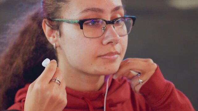 Young woman wearing glasses adjusting earphone in soft indoor light red hoodie closeup montage shows student inserting earbuds, commuter checking fit, podcast listener pausing to tune sound, attentive