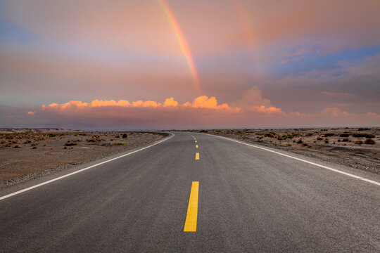 Empty asphalt road stretching through the desert under a vibrant double rainbow at sunset