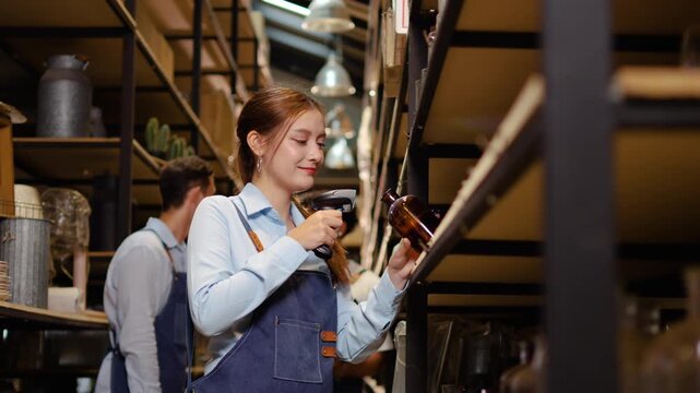 Smiling Asian female retail worker in a blue apron scanning product inventory with a barcode reader on a warehouse shelf. She checks store stock, then turns to look and smile at the camera happily.