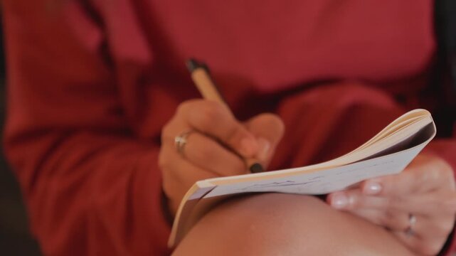 Woman scribbling in notebook under city lights, evening bokeh and neon glow, candid street mood on a bench, hoodie sleeve and rings visible, pen tracing notes in cinematic grain, solitary reflective