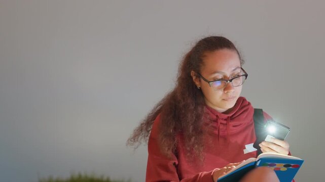 City skyline reading under phone light, red hoodie student holding flashlight, illuminated pages, distant tower lights, concentrated study, checking map and notes while skyline glows