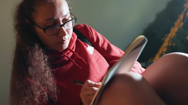 Person writing in notebook with glasses, red sweater and long curly hair, concentrated expression, indoor warm light, journal on lap, thoughtful composition, studying or drafting, candid portrait