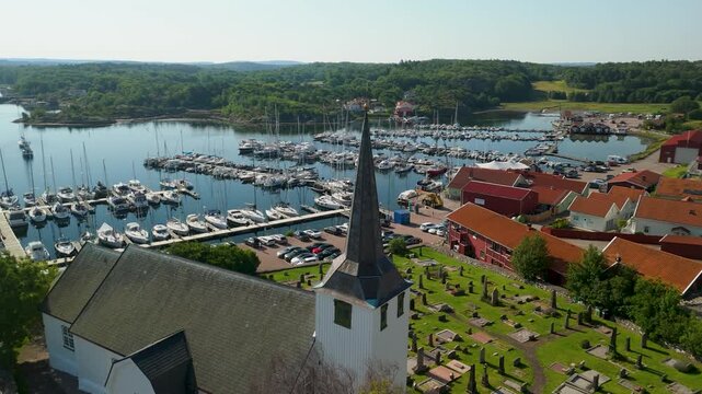 Aerial view of Fiskeb&auml;ckskil white church and cemetery beside bustling marina in Bohusl&auml;n Sweden with sailboats filling calm sheltered inlet surrounded by forest - orbit shot