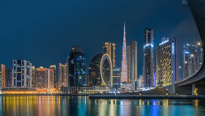 Fototapete Skyline Modern buildings in the city center of Dubai in the evening. Illuminated skyscrapers in the financial center at the blue hour. View of skyline in united arab emirates at night and reflection   © Mina