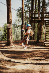 Fototapeta premium A girl is soaring through the air on a zip line, her face radiating joy and excitement. Dressed in a helmet and gloves, she is surrounded by tall trees and the adventurous spirit of the rope park.
