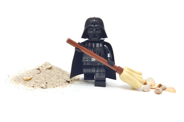 Naklejka premium Lego minifigure of peaceful Darth Vader from Star Wars is cleaning a sand from seashell isolated on white. Editorial illustrative image of stop war concept.