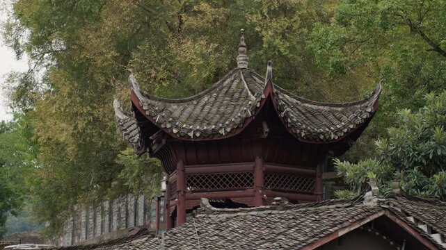 Ancient wooden pavilion roof under trees, weathered tile eaves curling skyward with carved timber beams, mosscovered ridge and stone wall, lush foliage and soft autumn light creating serene heritage