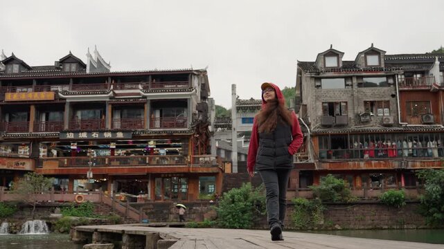 Woman walking river historic wooden buildings, backpacker explores overcast heritage town with narrow docks, balconies, stone steps, market stalls and calm water reflections, moody solitary atmosphere