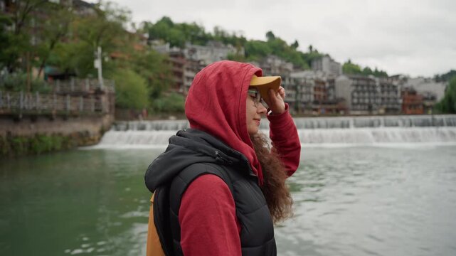 Caucasian woman in red hoodie riverside, profile view wearing cap and backpack, facing river dam and row of historic buildings under overcast sky, contemplative expression, travel mood, cool color