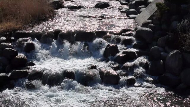 Water rushes over rocks in a wild, natural stream. Sunlight glints off the churning, frothy surface. Rocks line the streambed, creating a natural waterfall. Concept of untamed nature's power