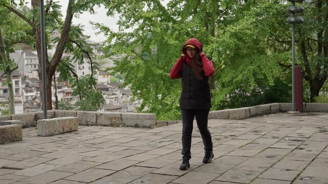 Frontal walker conversing on phone in plaza, red hoodie and yellow backpack facing camera, adjusting hood then speaking into mobile device first clip shows commuter checking directions while walking