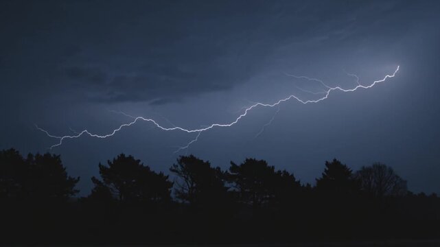 Powerful lightning strike illuminates dark stormy night sky over silhouetted trees