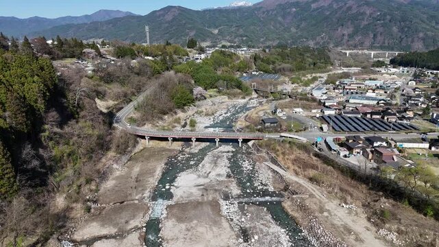 River winds through rugged terrain. Bridge spans the water's rushing current. Road curves along the hills' edge. Village lies nestled below the mountains. Sunlight bathes the scene in gentle light