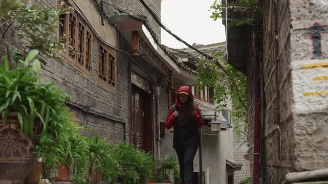 Woman walking down narrow stone alley, pausing to study carved windows and ivycovered walls, wearing red hoodie and black vest, slow contemplative stroll, soft overcast light, vintage architecture,
