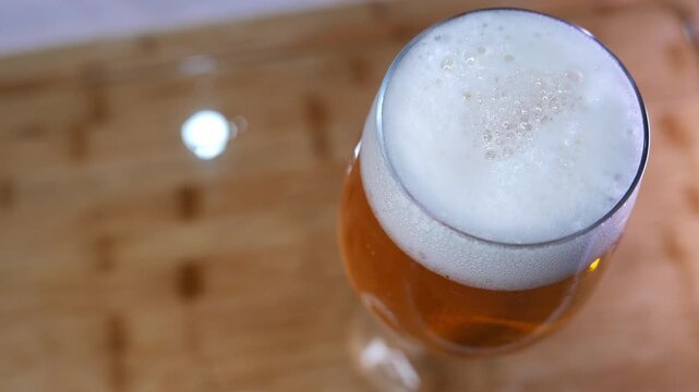 Zooming out from a beer glass on the table. Close-up shot of bubbles bursting in beer foam. A beer glass. A beer cup. A frothy drink. A beverage. A cold drink. A big beer head. Table top view.