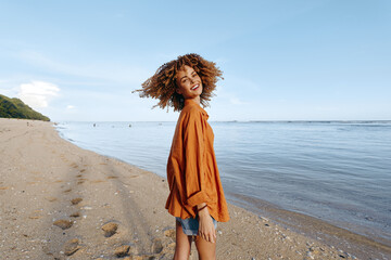 Young woman walking on sandy beach, smiling and looking back, casual outfit with orange shirt,...