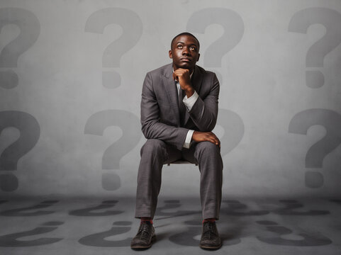 A focused young businessman in a suit sits contemplatively amidst many question marks on a grey background