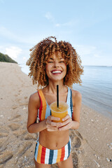Happy woman with curly hair wearing colorful swimsuit holds tropical drink with straw on sandy...