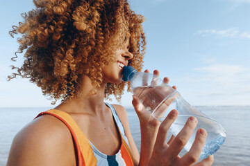 A smiling woman with curly hair drinks water from a plastic bottle outdoors near the sea. The woman...