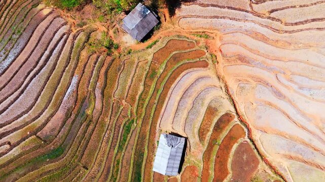 Layers of Vietnam hillside terraced water filled colourful brown rice paddies, prior to growing season. Curves from above with huts, drone lift reveal intricate patterns.