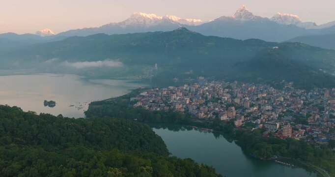 Aerial drone shot capturing golden sunrise over Phewa Lake Pokhara Nepal with Tal Barahi Temple surrounded by lush green hills and Annapurna and Machhapuchhre peaks in Lakeside with buildings.