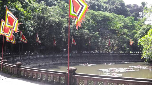 traditional Vietnamese five-color flags line stone railing beside quiet pond with water lilies under lush green mountains in Ninh Binh Vietnam.