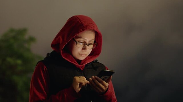 Outdoor night portrait person hoodie phone, park foliage and soft bokeh lights behind, focused browsing with thumb tapping screen, cautious walker checking map, cinematic grainy mood and cool evening