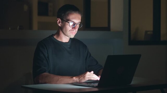 Freelancer in glasses working at night on laptop in dark room, focused man typing code and emails. Night work freelance job, remote work online, late night productivity and digital lifestyle at home