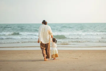 Rear view of a mother and her young daughter walking hand-in-hand towards the gentle sea waves on a sandy beach. Concept of family bonding, summer vacation, and wellness during a peaceful coastal trip © oatawa