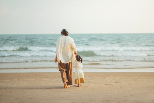 Rear view of a mother and her young daughter walking hand-in-hand towards the gentle sea waves on a sandy beach. Concept of family bonding, summer vacation, and wellness during a peaceful coastal trip