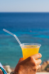 Close up of woman hand holding fresh orange cocktail in plastic cup with straw against blurred blue...