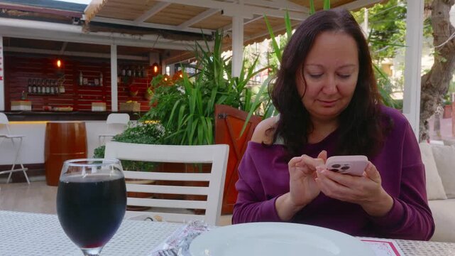 A mature woman with long dark hair sits alone at an outdoor restaurant table. She is focused on using her smartphone, scrolling and tapping. A glass of red wine is next to a white plate