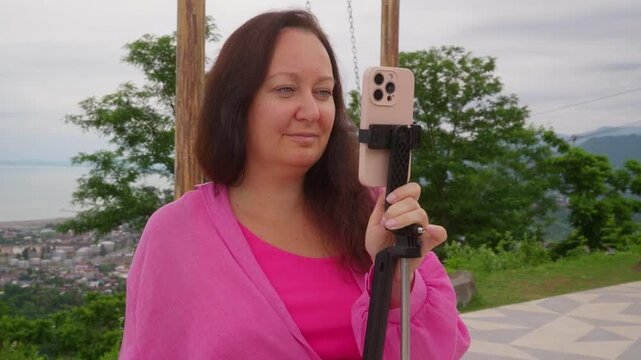 A brunette woman in pink clothes sets up her smartphone on a selfie stick to record a video or take a photo. She is at a scenic viewpoint overlooking the sea, a town, and mountains