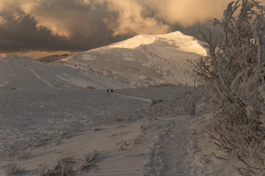 Epic winter panorama of Smerek peak in the Bieszczady Mountains with bright sunlight hitting the snowy summit against dark, dramatic storm clouds.