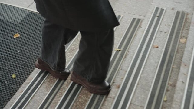 Brown shoes stepping on wet mat, man in long coat pauses on textured rubber entrance mat, slow deliberate steps, single yellow leaf, rainy urban morning, creator preparing to cross threshold into