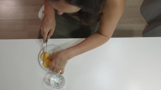 Top down view of a woman sitting at a white table. She uses a knife to slice a fresh peach on a plate. She then eats a piece of the fruit. A glass of water is also on the table