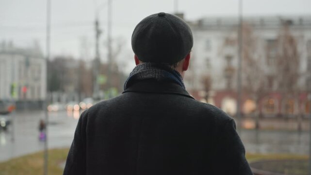 Back view white creator in city standing on wet plaza under overcast sky, wearing dark coat, beret and scarf, observing tramlines and muted architecture, mood of quiet introspection and street poet