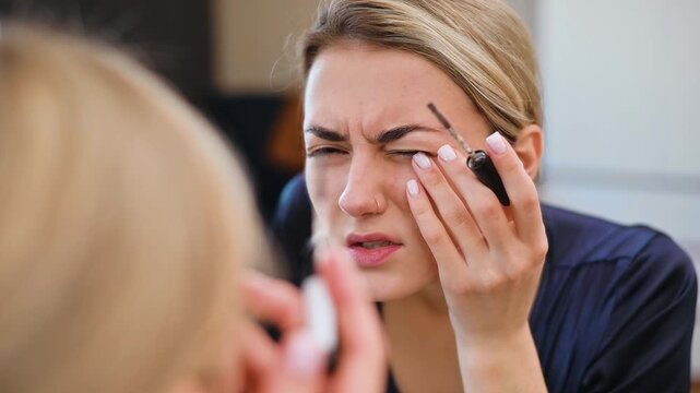 A woman applies mascara while looking into a bathroom mirror. The focus is on the makeup application process, showcasing beauty routines and techniques