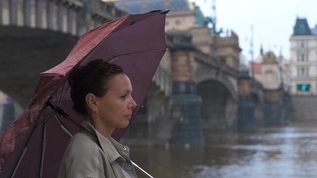 Lonely tourist with umbrella in the rain. Rain-drenched riverbank showcasing solitary tourist woman holding umbrella near vltava river's tranquil waters amid prague's atmospheric cityscape