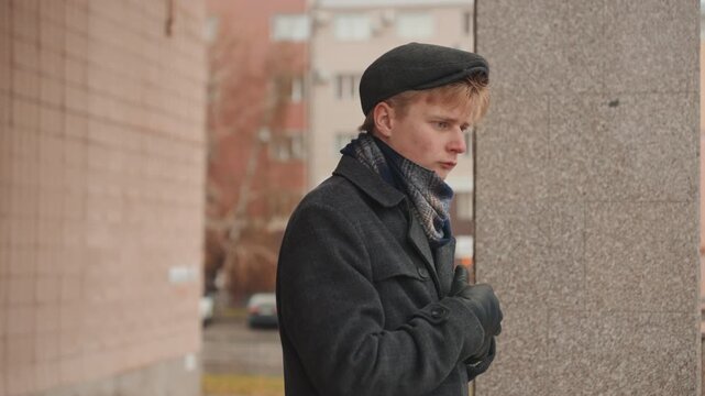 Pensive white man by column, wearing flat cap wool coat and gloves, leaning against textured concrete pillar, urban apartment blocks in background, muted overcast light, contemplative gaze and folded